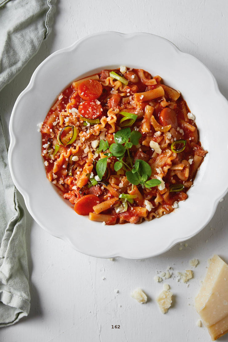 Tomatbasert pasterett i en dyp tallerk med ferske urter på toppen. Ved siden av tallerken ser vi en bit parmesanost. 
